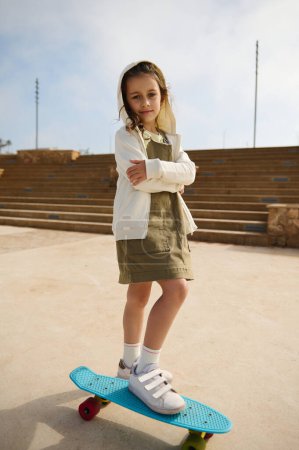 Téléchargez les photos : Enfant blanc adorable confiant fille, 6 ans, vêtue d'une robe kaki élégante et à capuche crémeuse, regardant la caméra, debout avec les bras pliés sur son skateboard sur une jambe sur l'aire de jeux urbaine - en image libre de droit