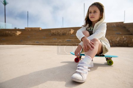 Téléchargez les photos : Portrait d'une adorable petite fille caucasienne regardant au loin tout en étant assise sur son skateboard à l'extérieur. Bonne enfance. Les gens. Loisirs. Mode de vie sain actif - en image libre de droit