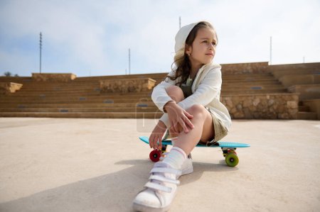 Téléchargez les photos : Adorable petite fille blanche assise sur son skateboard, regardant vers l'extérieur. Les gens. Enfance. Loisirs. - en image libre de droit