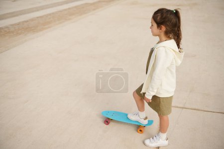 Téléchargez les photos : Vue aérienne de la petite fille blanche regardant loin tout en jouant au skateboard, ou le surf skate et commencer sur la piste de pompe extérieure dans le skatepark par des sports extrêmes. Les gens. Enfance. Loisirs - en image libre de droit