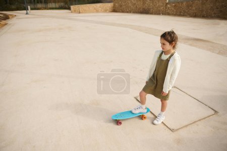 Téléchargez les photos : Vue d'en haut de la belle petite fille blanche debout sur son skateboard sur une jambe, regardant au loin, debout seul sur l'aire de jeux skatepark. Enfance. Loisirs - en image libre de droit