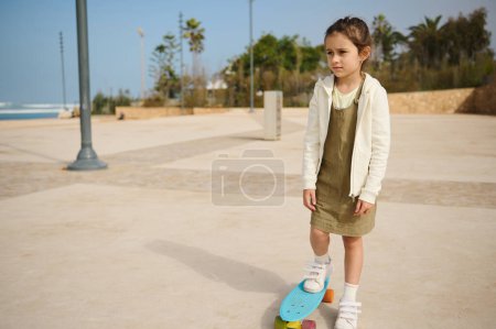Téléchargez les photos : Enfant caucasien mignon fille réfléchie rêvant dans la distance, debout sur son skateboard bleu sur une jambe sur un skate park urbain moderne. Bonne enfance. Concept de mode de vie actif sain - en image libre de droit