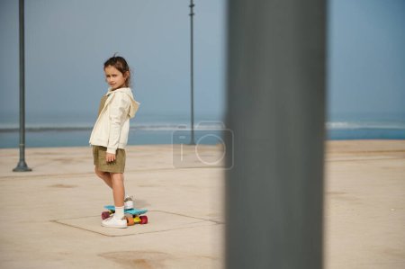 Téléchargez les photos : Adorable petite fille regardant la caméra, debout sur son skateboard bleu sur une jambe sur un skate park urbain moderne, sur fond d'océan. Bonne enfance. Concept de mode de vie actif sain - en image libre de droit