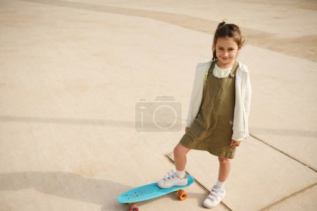 Téléchargez les photos : Souriant adorable petite fille debout sur un skateboard au skatepark urbain et regardant la caméra. Les gens. Mode de vie. Loisirs. Concept de divertissement pour enfants. Le sport extrême. Enfance - en image libre de droit