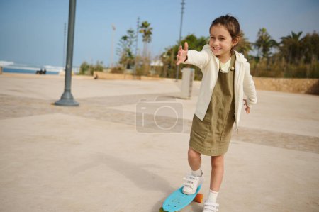 Téléchargez les photos : Heureux caucasien âge élémentaire enfant fille souriant mignonne, profiter du skateboard, monter sur skateboard sur un skatepark par une belle journée ensoleillée. Enfance. Concept de sport extrême - en image libre de droit