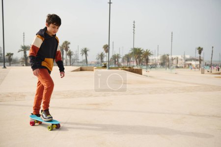 Téléchargez les photos : Heureux garçon d'âge élémentaire caucasien appréciant le skateboard, chevauchant sur le skateboard sur un skatepark par une belle journée ensoleillée. Enfance. Concept de sport extrême. Mode de vie sain et actif. Loisirs - en image libre de droit