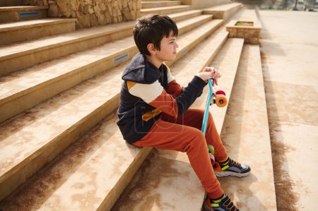 Téléchargez les photos : Portrait d'un adolescent, écolier en tenue de sport assis sur les marches du skatepark urbain, regardant attentivement au loin. Skateboard d'été, mode de vie actif. Loisirs sportifs - en image libre de droit