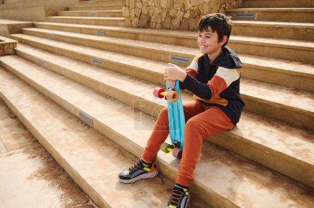Téléchargez les photos : Portrait horizontal complet d'un adolescent multi-ethnique en vêtements de sport, tenant son skateboard bleu, souriant, assis sur des marches à l'extérieur. Les gens. Du sport. Copier l'espace publicitaire pour le texte - en image libre de droit
