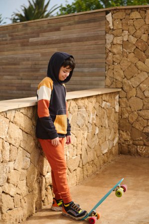 Téléchargez les photos : Portrait complet d'un enfant garçon heureux dans des vêtements de sport élégants, mettant une jambe sur sa planche de penny, souriant en regardant vers le bas, debout contre un fond de mur de pierre à l'extérieur. Loisirs. Les gens - en image libre de droit