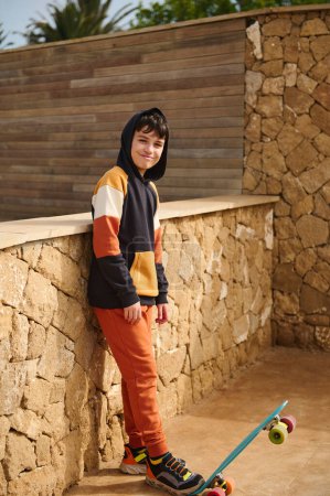Téléchargez les photos : Portrait complet d'un adolescent beau garçon en vêtements de sport, jouant avec une planche à roulettes, souriant à la caméra, debout à l'extérieur sur un fond de mur de pierre. Les gens. Du sport. Mode de vie sain actif - en image libre de droit
