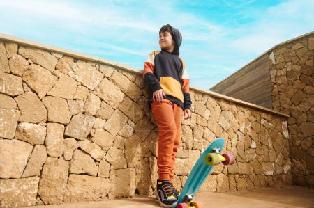 Téléchargez les photos : Portrait complet d'un adolescent beau garçon en vêtements de sport, jouant avec le skateboard, regardant de côté, debout à l'extérieur sur un fond de mur de pierre. Les gens. Du sport. Mode de vie sain actif - en image libre de droit
