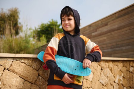 Téléchargez les photos : Portrait style de vie d'un adolescent, hipster, skateboarder en sweat à capuche tendance, tenant son skateboard bleu, souriant en regardant loin, debout à l'extérieur, sur un fond de mur de pierre. Copier l'espace publicitaire - en image libre de droit