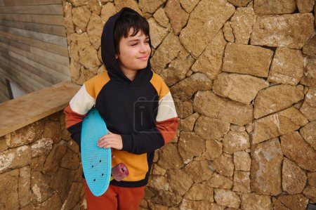 Téléchargez les photos : Portrait style de vie d'adolescent latino-américain beau garçon, hipster, skateboarder en sweat à capuche à la mode, tenant skateboard bleu, souriant regardant de côté un espace de copie, debout sur un fond de mur de pierre - en image libre de droit