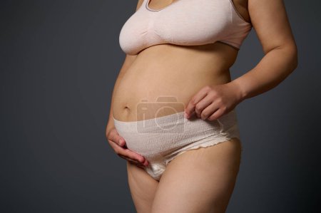 Photo for Close-up young mother holding her postnatal belly, showing stretch marks and her tummy after childbirth, standing isolated over gray studio background, dressed in maternity underwear. Body positivity - Royalty Free Image