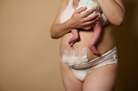 Cropped view of a body positive woman, new mother standing together with her newborn baby. Real body of women after few days of childbirth, isolated on beige studio background. Copy advertising space