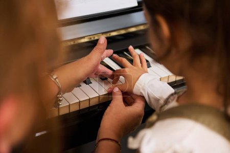 Close-up rear view from above of a little child girl learning playing piano, touching piano keys under the guidance of her teacher during individual music lesson indoor. Art, culture and entertainment