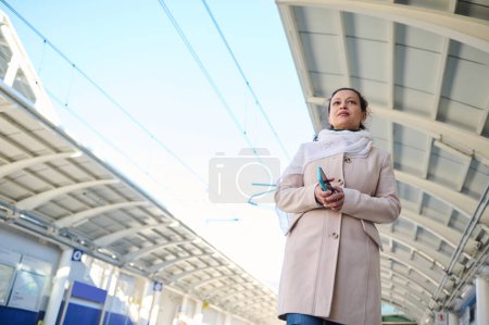 View from the bottom of a young happy woman commuter holding smart mobile phone, waiting to board the train, standing on the platform of a railway station, against blue sky background on winter day