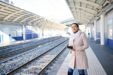 Attractive young brunette woman, business lady, commuter in beige coat, waiting to board the train on the railway station, standing on the platform with a smart phone in hands. People. Railroad trip