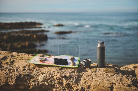 Photo for Still life with navigational equipment and thermos flask on the cliff against Atlantic ocean background while waves splashing while breaking on the headland. Compass, binoculars on the tourists map - Royalty Free Image