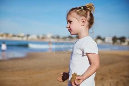 Side portrait of a beautiful Caucasian little child girl holding a wet sand while playing on the beach, thoughtfully looking into the distance. Happy carefree childhood concept
