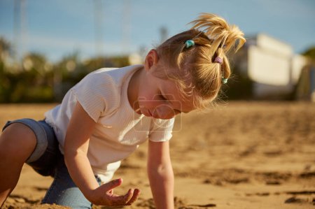 Entzückende kaukasische Blondine, die mit nassem Sand am Strand spielt, barfuß steht und am nassen Sandstrand Fußspuren hinterlässt. Menschen. Ein aktiver gesunder Lebensstil. Glückliche unbeschwerte Kindheit