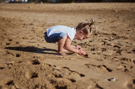 Entzückendes kleines Mädchen, das mit nassem Sand am Strand spielt, barfuß steht und am nassen Sandstrand Fußspuren hinterlässt. Menschen. Ein aktiver gesunder Lebensstil. Glückliche unbeschwerte Kindheit