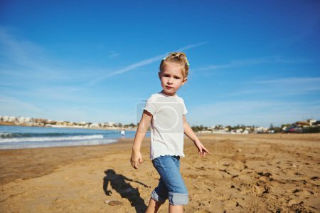 Entzückende kaukasische blonde Kindermädchen in weißem T-Shirt und blauer Jeans, die barfuß durch den nassen Sand in die Kamera schaut und ein glückliches Wochenende mit ihrer Familie im Freien genießt.