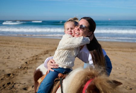 Téléchargez les photos : Adorable petite fille blanche embrassant sa mère tout en montant à cheval sur la plage. Mignon bébé fille apprendre l'équitation, assis sur un poney tout en appréciant le temps heureux avec sa maman aimante en plein air - en image libre de droit