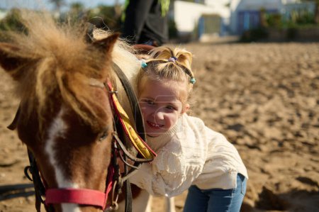 Téléchargez les photos : Adorable enfant fille embrassant doucement son cheval de poney, debout sur la plage de sable fin. Le concept de la thérapie alternative du cheval. Amour, soins et gentillesse pour les animaux. Heureuse enfance insouciante - en image libre de droit