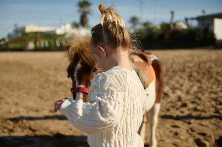 Téléchargez les photos : Vue arrière d'une petite fille blanche avec un poney sur la plage de sable - en image libre de droit