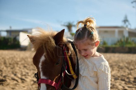 Téléchargez les photos : Adorable petite fille blonde caressant un poney, souriant et profitant d'un week-end heureux en plein air. Enfants et amour pour les animaux - en image libre de droit