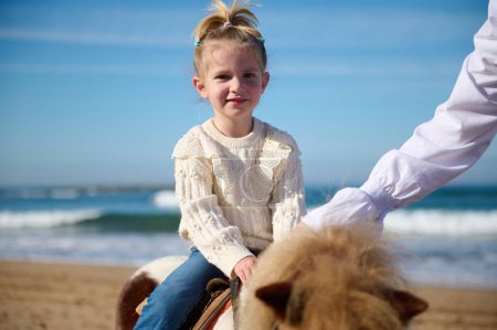 Téléchargez les photos : Heureuse petite fille caucasienne souriante regardant la caméra, chevauchant un cheval poney sur la plage, souriant regardant la caméra. Les gens. Mode de vie sain et actif. Enfants et animaux. activités de loisirs - en image libre de droit