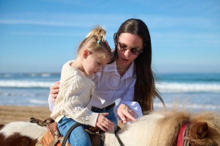 Téléchargez les photos : Une mère aimante et aimante qui apprend à sa fille à monter à cheval sur la plage. Les gens. Mode de vie sain et actif. Heureuse enfance insouciante. Loisirs et loisirs - en image libre de droit