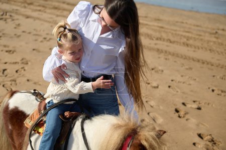 Téléchargez les photos : Petite fille blonde caucasienne se sentant inquiète et embrassant sa mère tout en chevauchant poney sur la plage de sable fin. Mon et ma fille profitent de moments heureux ensemble - en image libre de droit