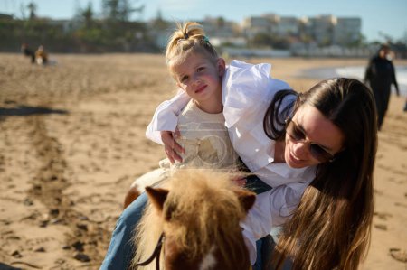 Téléchargez les photos : Mère souriante caressant un petit poney tout en soutenant sa fille sur l'apprentissage de l'équitation sur la plage de sable fin. Les gens. Mode de vie sain et actif. Loisirs et loisirs - en image libre de droit