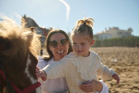 Téléchargez les photos : Mère heureuse portant sa fille tout en caressant un petit poney en plein air. Les gens. Animaux dans la nature sauvage - en image libre de droit