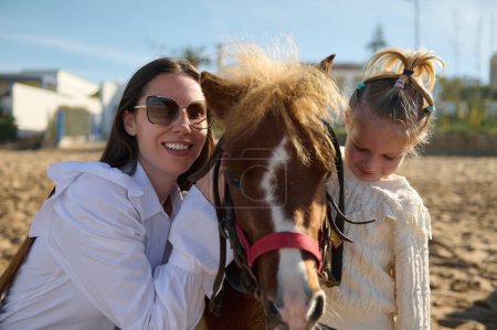 Téléchargez les photos : Joyeux jeune mère et fille souriant regardant la caméra, étreignant et posant avec un petit cheval de poney. Les gens. Loisirs et passe temps. Mode de vie sain actif - en image libre de droit