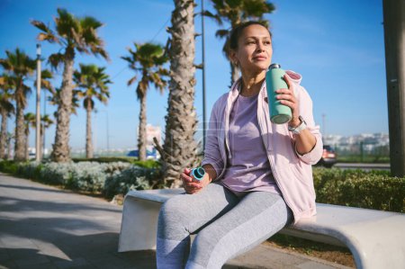 Attraktive sportliche Frau in Aktivkleidung, auf einer Bank an der Meerespromenade sitzend, nach dem Training oder Joggen erfrischendes Wasser trinkend, entspannt und verträumt in die Ferne blickend. Menschen und Sport