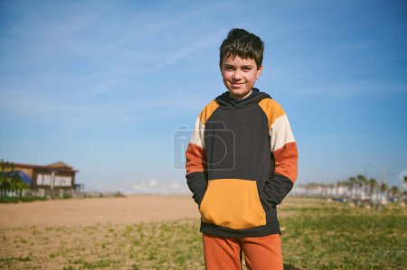 Confident authentic portrait of a multi ethnic handsome cheerful teenage school boy in sportswear, putting hands on his hoodie's pockets and smiling looking at the camera, standing on the sandy beach