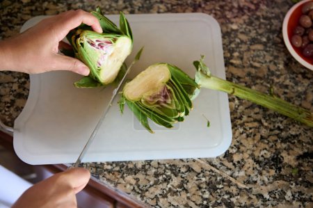 Foto de Vista desde arriba de las manos del chef cortando la flor de alcachofa en una tabla. Dos mitades de alcachofa orgánica fresca madura en la tabla de cortar - Imagen libre de derechos