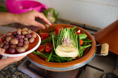 Foto de Primer plano ama de casa la adición de aceitunas en una comida mientras se cocina comida vegetariana saludable en tagine. Mujer preparando y sazonando verduras en casa - Imagen libre de derechos