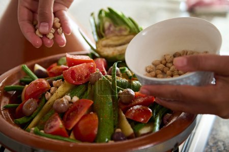 Foto de Primer plano de las manos de una ama de casa añadiendo garbanzos en una comida vegetariana mientras se cocinan verduras orgánicas saludables en maceta de tagine de arcilla. Fondo alimentario tradicional marroquí - Imagen libre de derechos