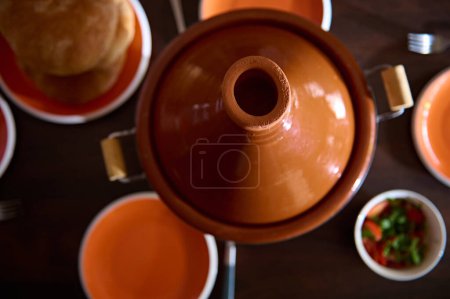 Foto de Tagine de maceta de arcilla vista superior, rotura recién horneada y platos de naranja limpios vacíos en la mesa de madera. Comida tradicional marroquí para la cena familiar - Imagen libre de derechos