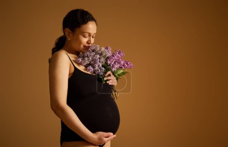 Confident studio portrait of a beautiful young pregnant woman in black bodysuit, holding her big belly in late pregnancy, posing with a bouquet of lilacs over beige background. Copy advertising space