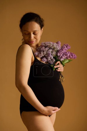Vertical studio shot of attractive delightful pregnant woman wearing black bodysuit, posing with a bunch of purple lilacs over beige background, enjoying her pregnancy time. Happy maternity concept