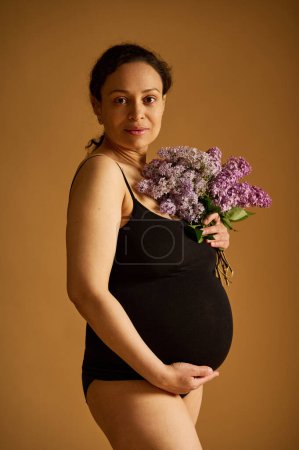 Confident multi ethnic body positive pregnant woman in black bodysuit, posing looking at the camera, isolated on beige studio background. Stretch marks. Pregnancy. Maternity lifestyle and childbering
