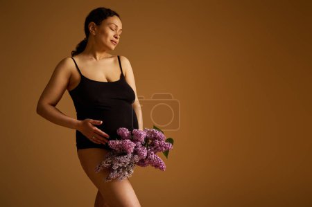 Authentic positive pregnant woman in black bodysuit, smiles while strokes her belly in late pregnancy, last trimester of happy carefree childbearing, posing over isolated beige studio background