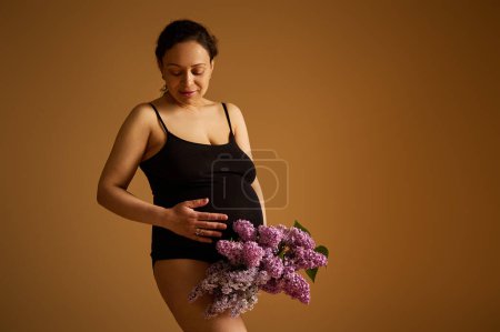 Portrait of a beautiful pregnant woman expecting baby, holding belly, looking aside, posing in black lingerie with a bunch of purple lilacs, isolated beige studio background. Pregnancy and maternity