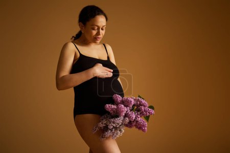 Authentic gravid female, future mother, pregnant woman in black bodysuit, holding lilacs, gently stroking her big belly, standing isolated on beige studio background. Happy carefree pregnancy concept