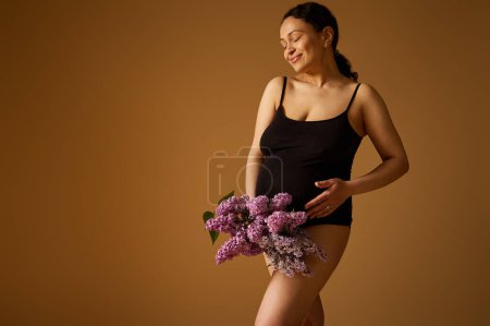 Side beauty portrait multi ethnic pretty pregnant gravid woman, expectant mother with big belly in pregnancy 28 week, wearing black lingerie, holding blooming lilacs, isolated beige studio background
